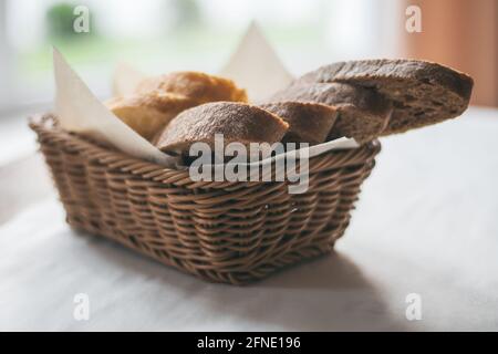 Teilweise verschwommener Korbkorb mit schwarzem und weißem Brot Weiße Tischdecke neben dem Fenster Stockfoto