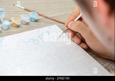 Eine mans Hand hält einen Pinsel und zeichnet ein Bild durch Zahlen. Zeichnen auf Leinwand. Nummerierte Farbdosen. Kreatives Hobby. Malen für Anfänger.Nahaufnahme, sele Stockfoto
