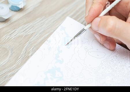 Eine mans Hand hält einen Pinsel und zeichnet ein Bild durch Zahlen. Zeichnen auf Leinwand. Nummerierte Farbdosen. Kreatives Hobby. Malen für Anfänger.Nahaufnahme, sele Stockfoto
