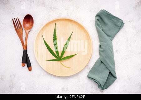 Essen konzeptuelles Bild eines Cannabisblattes mit Löffel und Gabel auf weißem Beton Hintergrund. Stockfoto