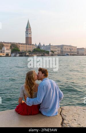 Junges verliebter Mann, der am Meer sitzt und küsst, auf dem hinteren Markusplatz mit Campanile di San Marco, Venedig, Venetien, Italien Stockfoto