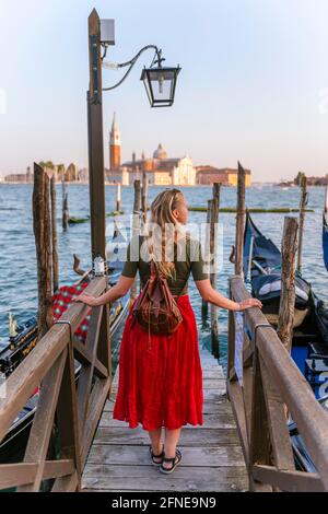 Junge Frau mit rotem Kleid auf einem Steg, venezianische Gondeln, in der hinteren Kirche San Giorgio Maggiore, Venedig, Venetien, Italien Stockfoto