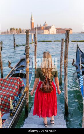 Junge Frau mit rotem Kleid auf einem Steg, venezianische Gondeln, in der hinteren Kirche San Giorgio Maggiore, Venedig, Venetien, Italien Stockfoto