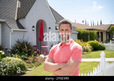 Lächelnder amerikanischer Mann mit gekreuzten Armen, der vor seinem neuen Zuhause steht. Kaufen, verkaufen, Immobilien, Immobilien, Hausversicherung Konzept. Echte Menschen. Stockfoto