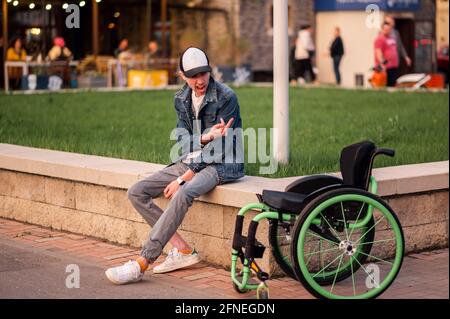 Ein junger behinderter Mann sitzt neben einem Rollstuhl und zeigt ihr den Mittelfinger Stockfoto