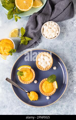 Minitörtchen mit Zitronenquark, Mini-Baiser, Zitronenscheiben und Minze auf blauem Betongrund. Draufsicht. Selektiver Fokus. Stockfoto