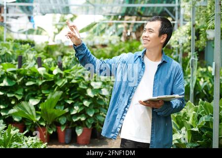 Glücklicher junger Mann, der gerne in seinem Gartencenter arbeitet, Pflanzen überprüft und sich Notizen in Dokumenten macht Stockfoto