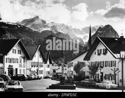 'Dieses undatierte Foto, aufgenommen um 1975, zeigt den Floriansplatz mit dem Floriansbrunnen in Partenkirchen, der Blick geht in die Ballengasse. An einer Ampel rechts hängt ein Schild mit der Aufschrift: ''zum öffentlichen WC 150 m''. Auf der linken Seite befindet sich ein Friseursalon. Rechts erhebt sich hinter den Häusern der Turm der Kirche Mariä Himmelfahrt. Im Hintergrund ragt das Wettersteingebirge mit dem Rießerkopf (vorne, Mitte des Bildes) und den schneebedeckten Gipfeln der Alpspitze, Höllentalspitzen, Zugspitze und Waxenstein (Hintergrund, Stockfoto