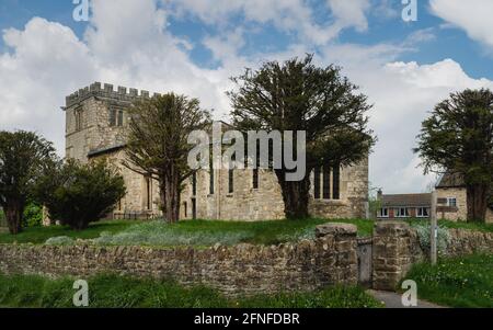 Alte Allerheiligen-Kirche, flankiert von Trockensteinmauern und Bäumen, die im Frühling unter einem wolkenlosen blauen Himmel stehen. Goodmanham, Yorkshire, Großbritannien. Stockfoto