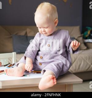 Entzückender kleiner Junge mit goldenem Haar in Leinenkleidung, der auf einem Holztisch sitzt und mit Buntstiften spielt, das Zeichnen lernt und Aktivitäten für Kleinkinder genießt. Frühkindliche Entwicklung Stockfoto