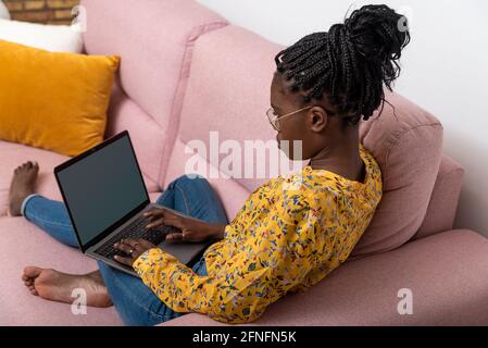 Hochwinklige Seitenansicht eines schwarzen Freiberuflers, der auf dem Sofa sitzt Und das Tippen auf Laptopund das Arbeiten auf dem Geschäft von zu Hause aus Stockfoto