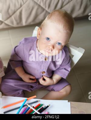 Süßes blauäugiges kleines Baby mit goldenem Haar in Leinenromper, das auf die Kamera schaut, während es am Schreibtisch sitzt und mit bunten Bleistiften zeichnet, Blick von oben. Frühkindliche Entwicklung Stockfoto