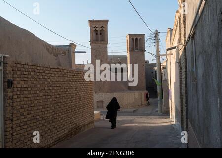 Frau in schwarzem Tschador, die in einer Straße mit einer ab-anbar, einem unterirdischen Wasserreservoir im Dorf Mohammadieh in der Nähe von Nain, Iran, läuft Stockfoto