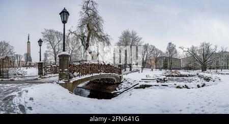 Blick auf den Bastion Hill Park und das Freiheitsdenkmal in Riga, Lettland. Winterlandschaft in verschneiten Park mit schöner kleiner Brücke mit Schleusen über Teich. Stockfoto