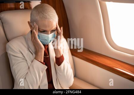 Geschäftsfrau in medizinischer Maske, die an Kopfschmerzen im Flugzeug leidet Stockfoto