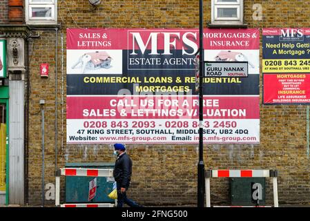 Ein asiatischer Mann mit Turban und Maske, der an einem vorbeiläuft Großes Straßenplakat, das einen lokalen Immobilienmakler in Southall wirbt London, England, Großbritannien Stockfoto