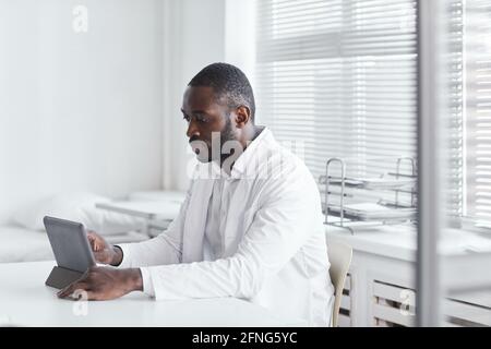 Afrikanischer junger Arzt sitzt am Tisch und arbeitet daran Digitales Tablet in seinem Büro Stockfoto