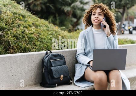 Fokussierte junge afroamerikanische Frau in blauem Mantel an der Arbeit Netbook und Telefonieren auf dem Mobiltelefon, während Sie auf Stein sitzen Bank im Stadtpark auf klaren s Stockfoto