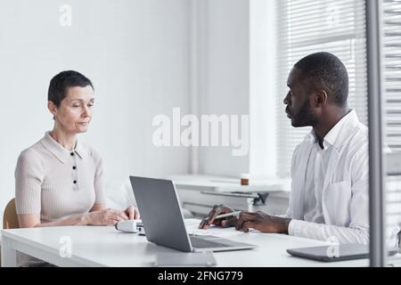 Afrikanischer Arzt sitzt am Tisch und spricht mit dem Patientin während ihres Besuchs in der Arztpraxis Stockfoto