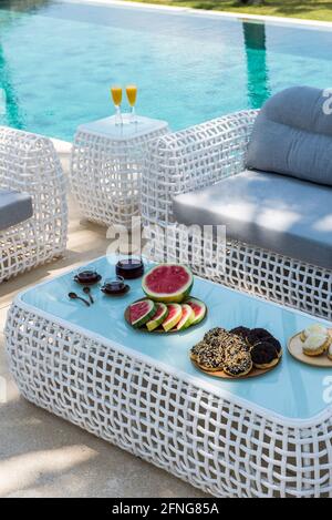 Von oben Teller mit verschiedenen appetitlichen Gebäck und frisch In Scheiben geschnittene Wassermelone auf dem Tisch serviert mit Tassen Kaffee in der Nähe Rattansofa am Pool Stockfoto