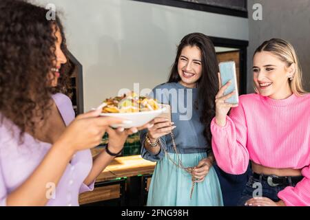 Fröhliche junge stilvolle Damen lächeln beim Fotografieren von Getreide Ethnische Freundin steht mit einem Teller appetitlich gemischter Früchte Salat Stockfoto
