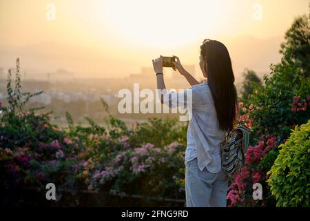 Ethnische asiatische Frau, die im Garten steht und während der Sonnenuntergangszeit mit dem Smartphone fotografiert Stockfoto