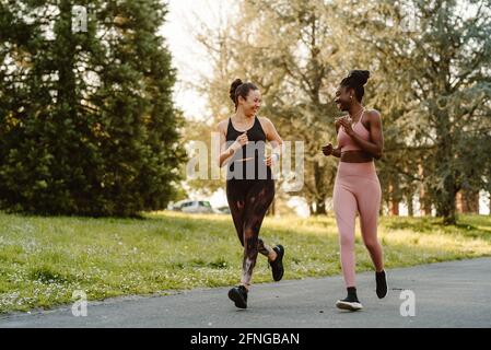 Lächelnde, multirassische Läuferinnen beim Joggen und Sprechen im aktiven Ohr Ausdauertraining auf Gehwegen in der Stadt Stockfoto