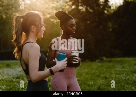 Junge, multirassische Sportlerinnen in Sportkleidung mit Getränken, die während des Gesprächs reden Blick auf den Rasen im Sonnenlicht Stockfoto