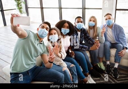 Gruppe multiethnischer Freunde in Gesichtsmasken, die Selfie mit dem Smartphone auf sozialen Treffen zu Hause machen und dabei Schutz tragen Stockfoto