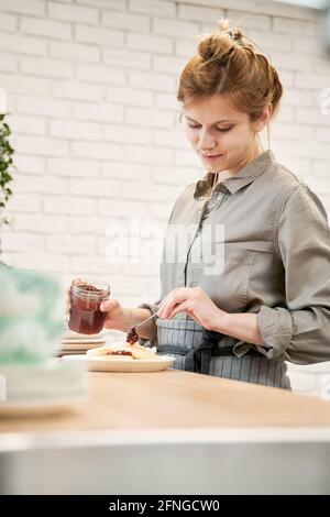 Junge Frau mit Krug, die köstliche Beerengelee auf Pfannkuchen setzt Beim Kochen am Tisch in der Küche Stockfoto