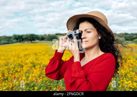 Lächelndes Weibchen im Hut, das mit einer alten Kamera fotografiert Wiese unter bewölktem Himmel Stockfoto