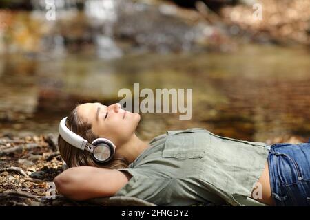 Seitenansicht Porträt einer glücklichen Frau, die sich liegend ausruht und zuhört Zu Musik mit kabellosen Kopfhörern am Flussufer Stockfoto