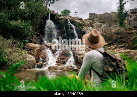 Rückansicht anonymer männlicher Rucksacktourist im Hut, der die Aussicht genießt Kaskade aus rauem Fels in grüner Natur Stockfoto