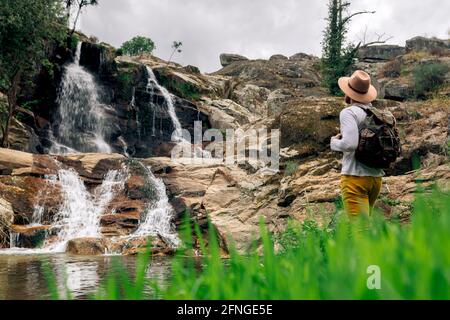 Rückansicht anonymer männlicher Rucksacktourist im Hut, der die Aussicht genießt Kaskade aus rauem Fels in grüner Natur Stockfoto