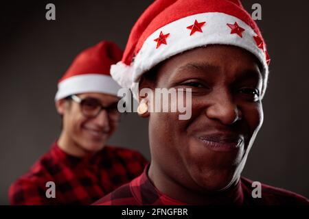 Charismatische männliche Freunde grinsen an der Kamera in Santa Hats Während sie die Weihnachtsfeiertage mit Fokus feiern An einen lächelnden schwarzen Mann Stockfoto