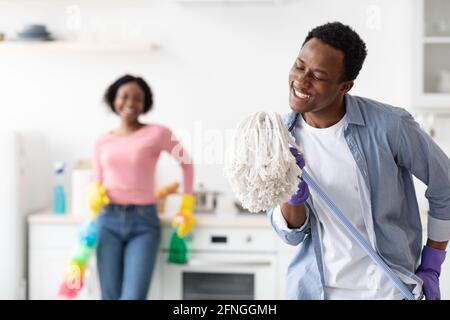 Fröhlicher schwarzer Kerl, der Mopp als Mikrofon verwendet und beim Reinigen singt Stockfoto