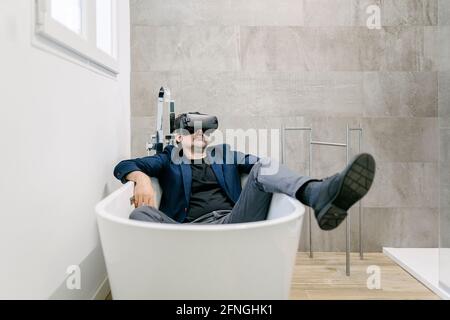 Vollständig bekleideter, eleganter erwachsener Mann, der in einer Badewanne virtuelle Realität erlebt, als wäre er an einem tropischen Strand. Virtual-Reality-Brille. Virtuelle Realität Stockfoto