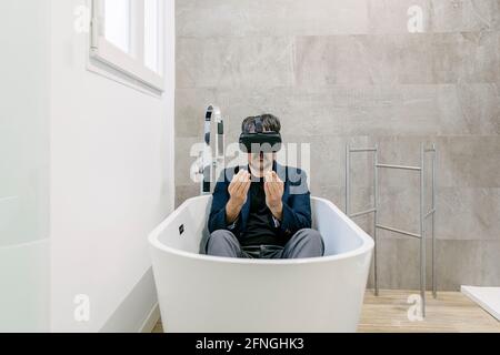 Vollständig bekleideter, eleganter erwachsener Mann, der in einer Badewanne virtuelle Realität erlebt, als wäre er an einem tropischen Strand. Virtual-Reality-Brille. Virtuelle Realität Stockfoto