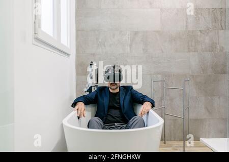 Vollständig bekleideter, eleganter erwachsener Mann, der in einer Badewanne virtuelle Realität erlebt, als wäre er an einem tropischen Strand. Virtual-Reality-Brille. Virtuelle Realität Stockfoto