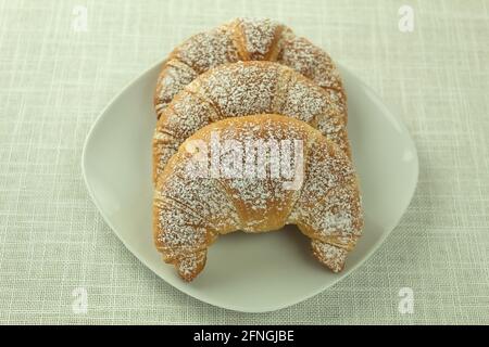 Croissants mit Puderzucker auf einem weißen Teller. Drei knusprige Croissants mit weißem Puderzucker auf einem weißen Teller. Hintergrund, Hintergrundbild Stockfoto