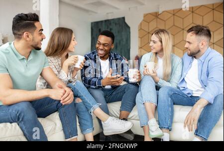 Gruppe von fröhlichen multiethnischen Freunden, die zu Hause feiern, auf der Couch sitzen und Kaffee trinken Stockfoto