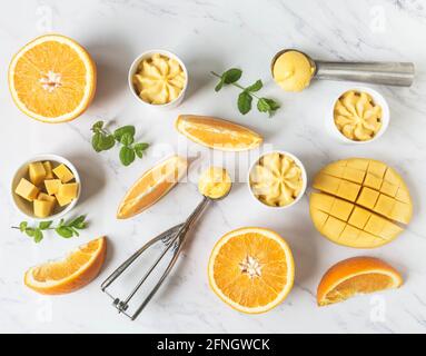 Orangen- und Mango-Sorbet in Schale und in Eislöffeln und Früchten Gewürze auf Marmorboden. Kalter Sommerdessert. Draufsicht, flach liegend Stockfoto