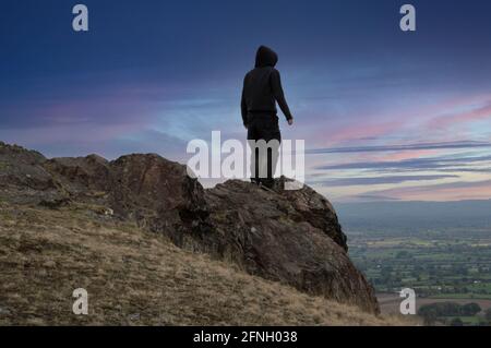 Ein launischer Mann mit Kapuze, zurück zur Kamera, der auf einem Berg steht und einen wunderschönen Sonnenuntergang anschaut. Stockfoto