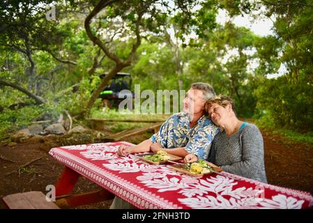 Verheiratetes Paar, das am Picknicktisch sitzt und ein romantisches Picknick-Abendessen auf einem Hügel auf der Gunstock Ranch bei Sonnenuntergang, Laie, Oahu, Hawaii, USA, genießt Stockfoto