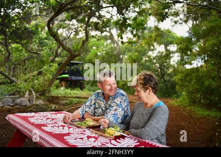 Verheiratetes Paar, das am Picknicktisch sitzt und ein romantisches Picknick-Abendessen auf einem Hügel auf der Gunstock Ranch bei Sonnenuntergang, Laie, Oahu, Hawaii, USA, genießt Stockfoto