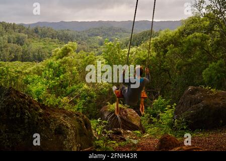 Frau, die auf einer Seilschaukel am Baum sitzt, auf dem Hügel der Gunstock Ranch, bei Sonnenuntergang, Laie, Oahu, Hawaii, USA Stockfoto