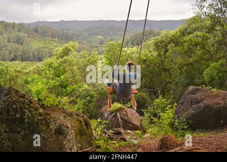 Frau, die auf einer Seilschaukel am Baum sitzt, auf dem Hügel der Gunstock Ranch, bei Sonnenuntergang, Laie, Oahu, Hawaii, USA Stockfoto