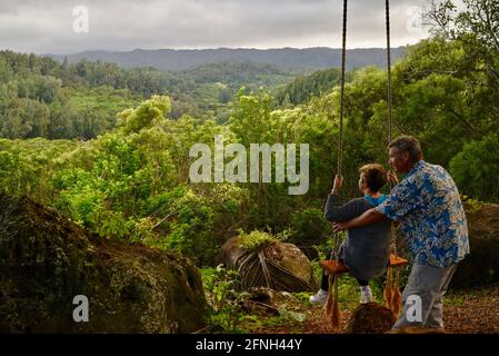 Verheiratetes Paar, Ehemann, der seine Frau auf einer Seilschaukel, die am Baum hängt, auf dem Hügel der Gunstock Ranch bei Sonnenuntergang, Laie, Oahu, Hawaii, USA, schiebt Stockfoto