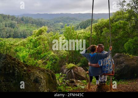 Verheiratetes Paar, Ehemann, der seine Frau auf einer Seilschaukel, die am Baum hängt, auf dem Hügel der Gunstock Ranch bei Sonnenuntergang, Laie, Oahu, Hawaii, USA, schiebt Stockfoto