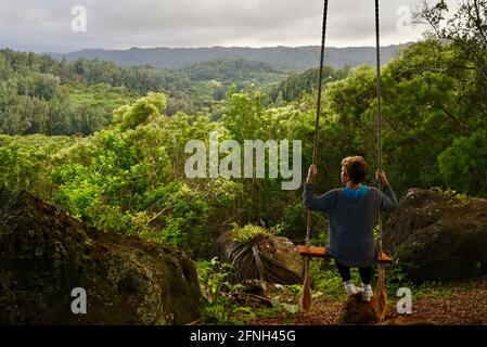 Frau, die auf einer Seilschaukel am Baum sitzt, auf dem Hügel der Gunstock Ranch, bei Sonnenuntergang, Laie, Oahu, Hawaii, USA Stockfoto